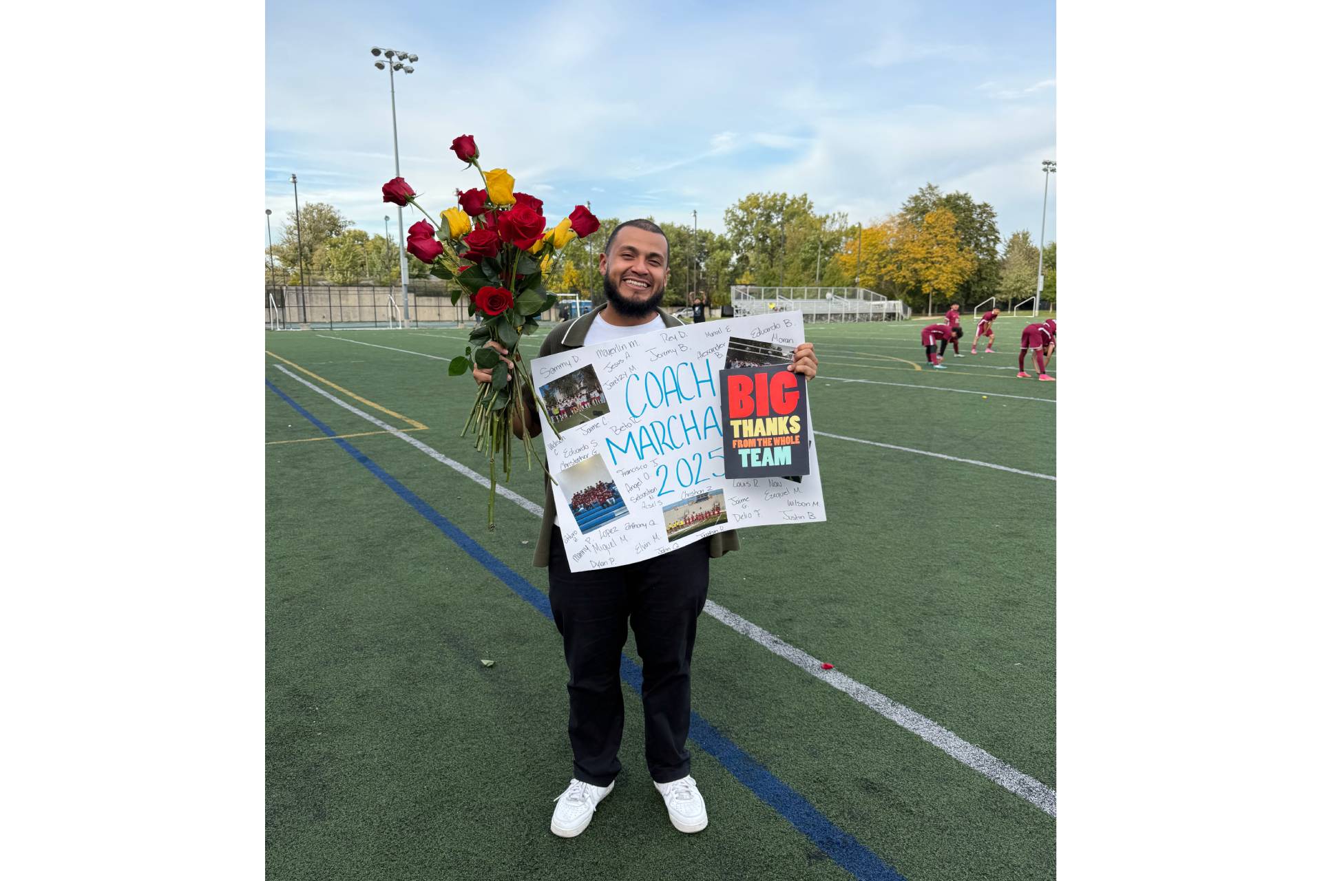 Froilan Marchan Jr., a social worker and soccer coach at North-Grand High School, smiling on the soccer field while holding a bouquet of flowers and a poster.